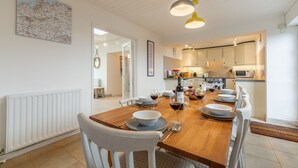 Un comedor con una mesa de madera dispuesta para seis personas, sillas blancas y una cocina al fondo.