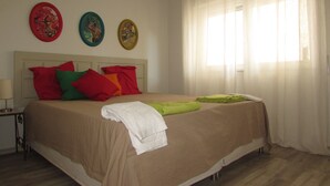 Una cama bien tendida con una colcha color crema y varias almohadas en rojo, verde y naranja. Encima de la cama hay tres adornos de pared, y una ventana con cortinas translúcidas que dejan entrar luz natural.