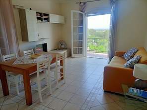 Un salon comprenant un canapé, une table à manger avec des chaises, un téléviseur, un ventilateur et une vue sur la verdure, visible à travers une porte-fenêtre donnant sur un balcon.