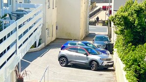 A narrow street with parked cars, a staircase leading to a building, and a view of the sea in the background.