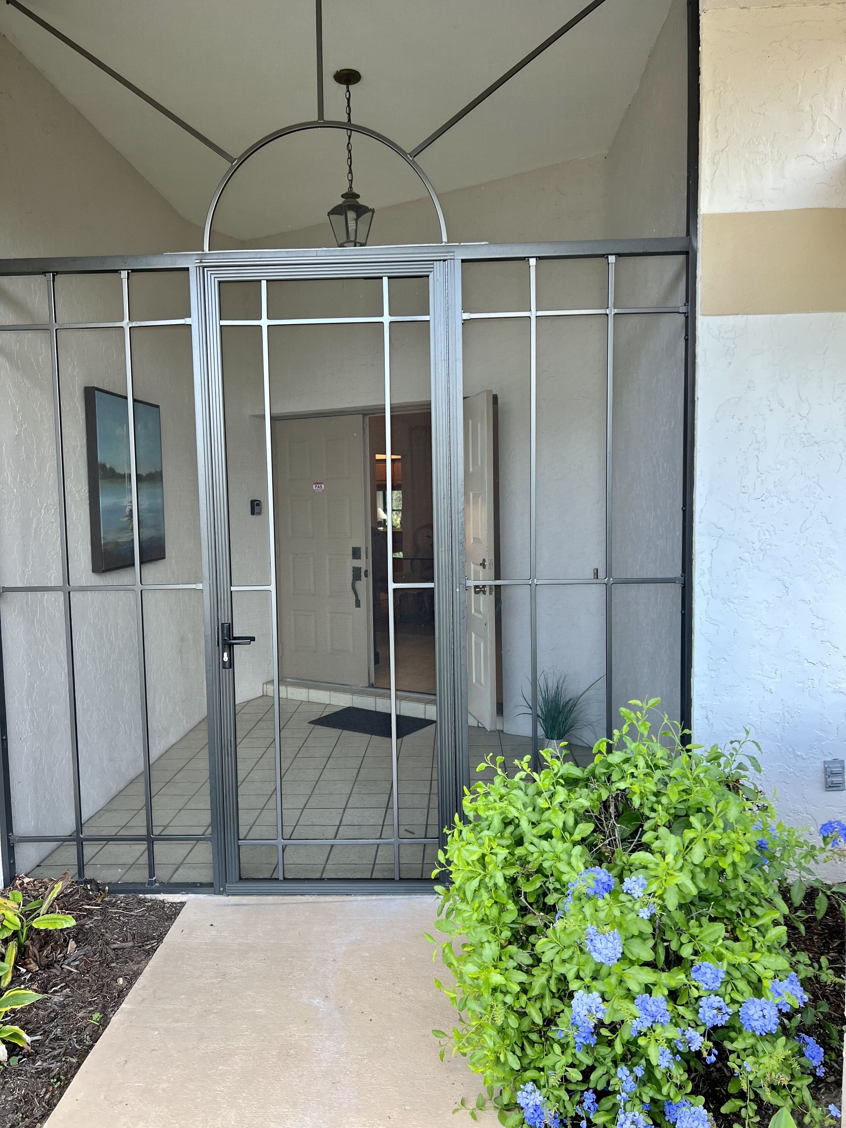 A gated entrance with a metal gate, a potted plant with blue flowers, and a tiled floor leading to an interior space.