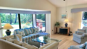 Un salon spacieux avec un canapé d’angle, une table basse en verre et un téléviseur. De grandes fenêtres offrent une vue sur un jardin.