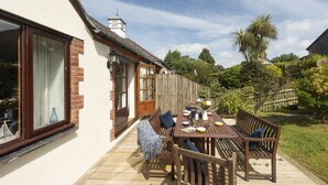 Une terrasse en bois avec une table et des chaises, une clôture en bois et un jardin avec des arbres et des arbustes.