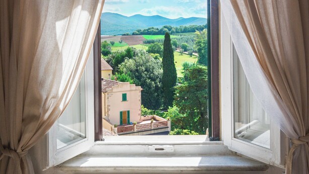 Una ventana con cortinas que muestra una vista de paisaje rural con campos verdes, árboles y montañas distantes.