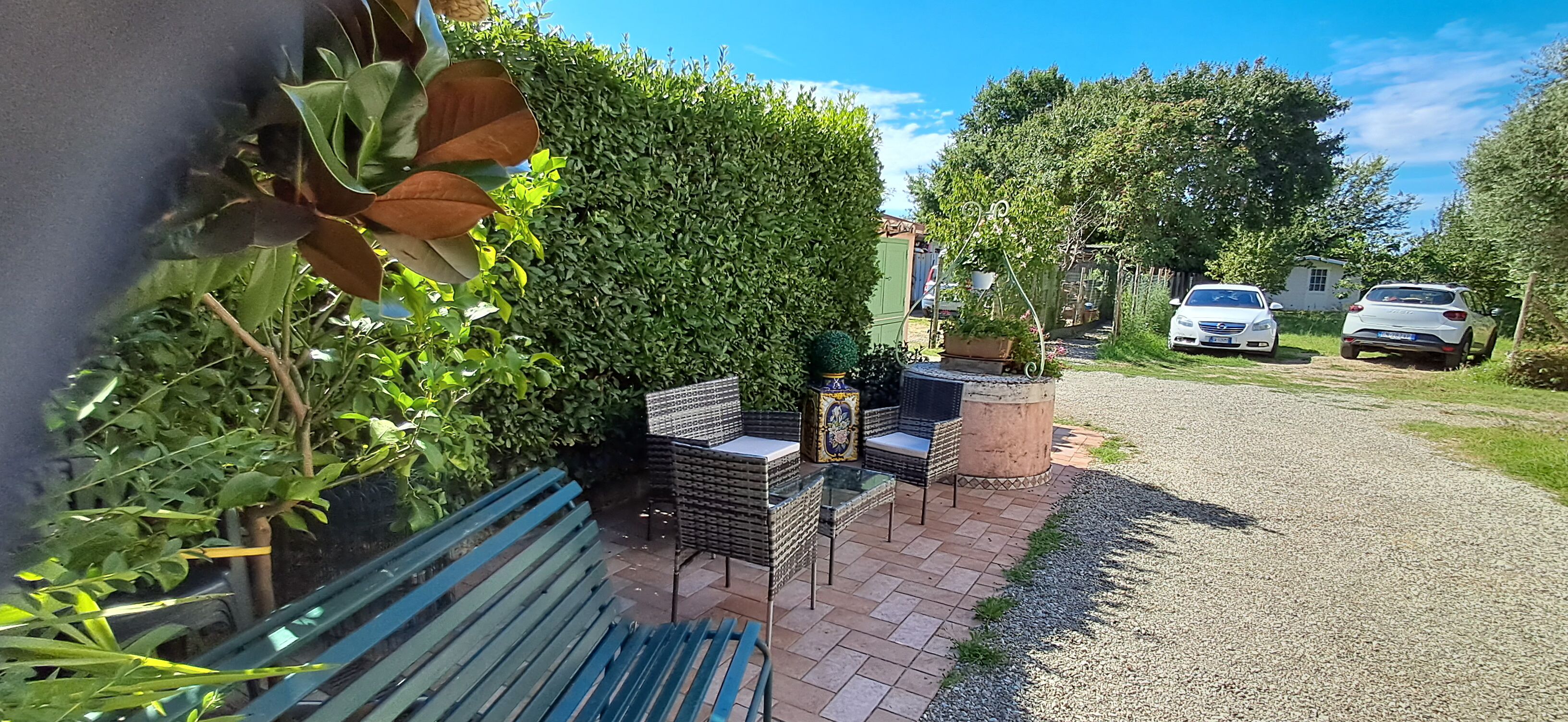 Eine Terrassenfläche mit einer Bank, Korbstühlen und einem Tisch, umgeben von Grünpflanzen und einem Hecken.