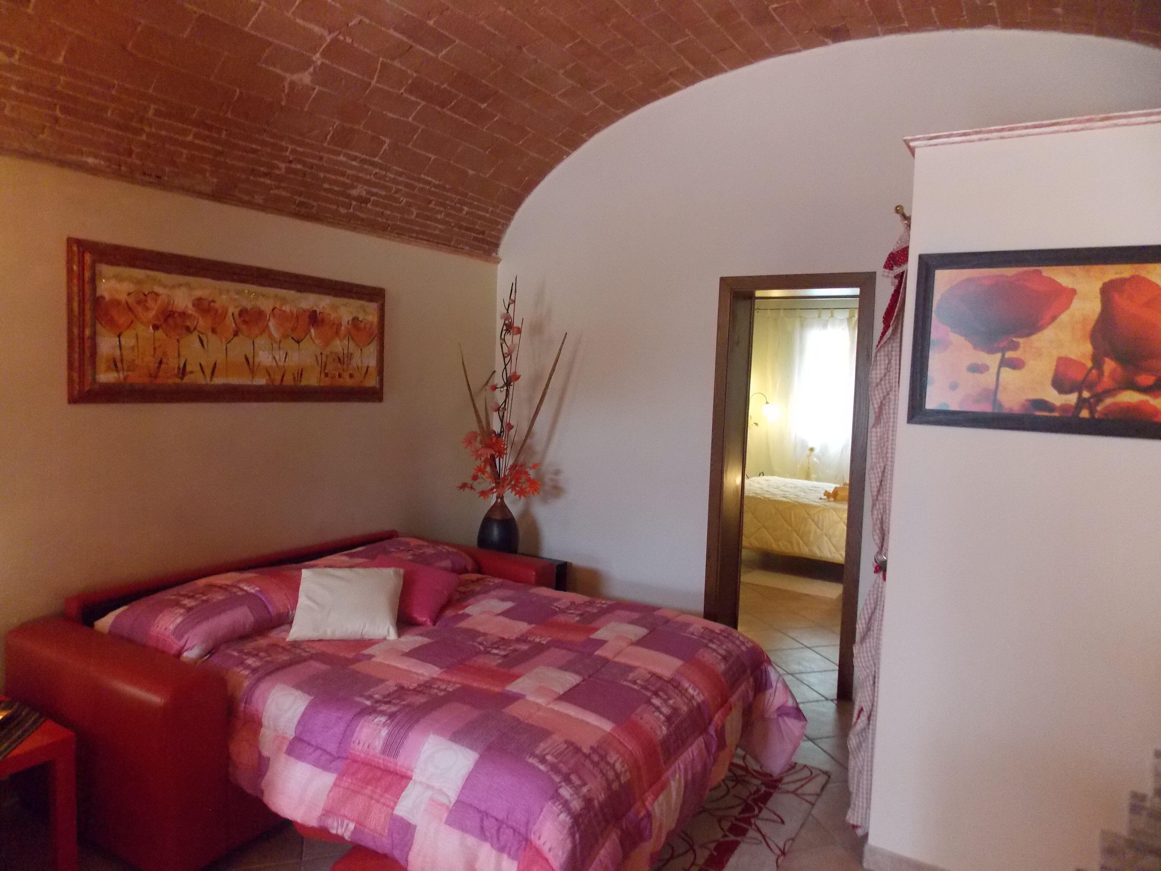 A bedroom with a brick ceiling, a bed with a pink and purple quilt, a vase with flowers, and a painting on the wall.