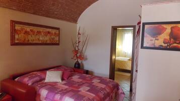 A bedroom with a brick ceiling, a bed with a pink and purple quilt, a vase with flowers, and a painting on the wall.