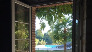 A sliding glass door leading to an outdoor patio area with a pool, a palm tree, and a wooden table and chairs. - Beautiful Cottage ****  on the bank of the river Tarn - 6/7 people. Pool . (Labastide de Levis)