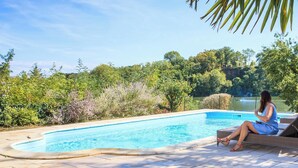 A woman sitting by a swimming pool with lounge chairs, surrounded by lush greenery and a clear sky. - Beautiful Cottage ****  on the bank of the river Tarn - 6/7 people. Pool . (Labastide de Levis)