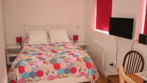 A bedroom with a bed, two bedside tables, a TV mounted on the wall, and a window with red curtains. - Self-contained New Studio with independent entrance in an up-market quiet area. (London)