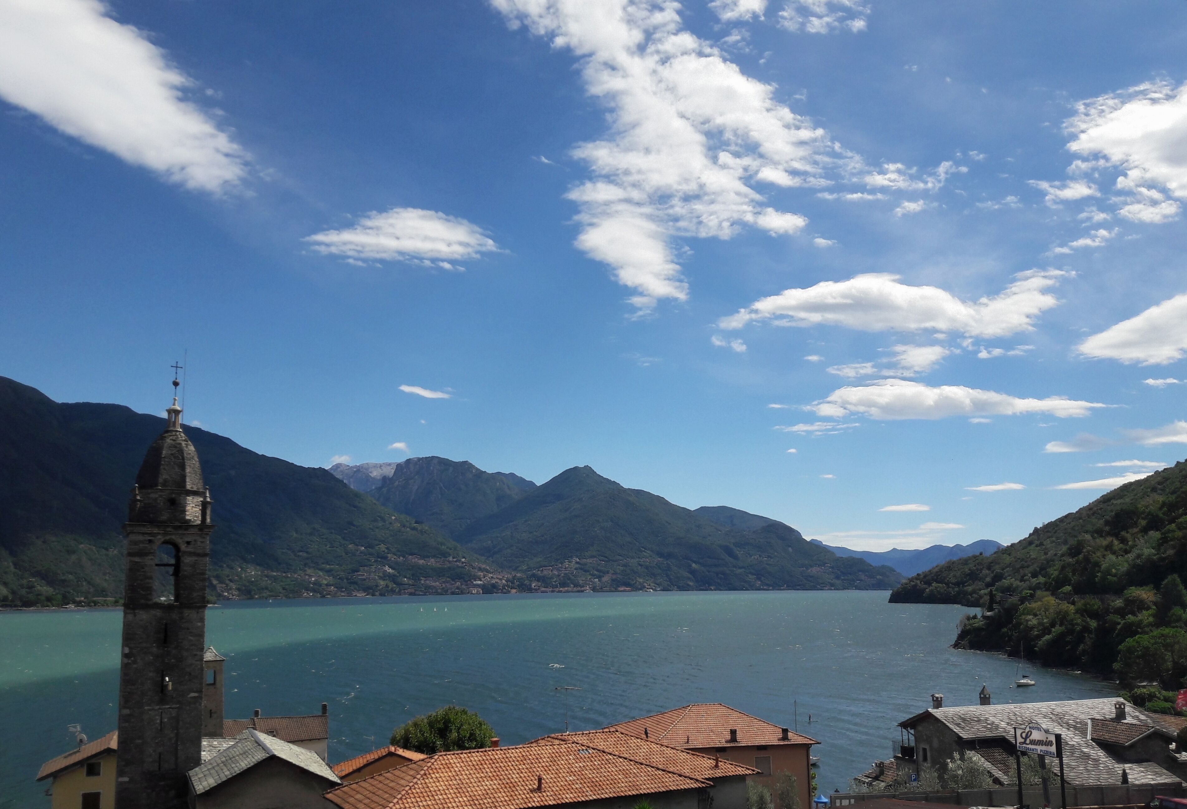 Vista de un lago con montañas al fondo, una iglesia con un campanario y un pequeño edificio con un cartel.