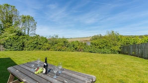 A wooden picnic table with a bottle and glasses, surrounded by a grassy yard and a wooden fence. - September is a lovely spacious house in the heart of Roserrow. (Wadebridge)