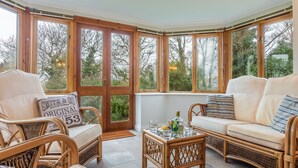 A corner room with wicker furniture, a glass-top table, and large windows offering a view of trees. - September is a lovely spacious house in the heart of Roserrow. (Wadebridge)