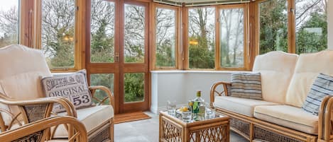 A corner room with wicker furniture, a glass-top table, and large windows offering a view of trees.