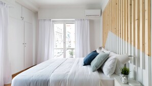 Quarto moderno com uma cama grande, cortinas brancas, painel de ripas de madeira e vista para edifícios.