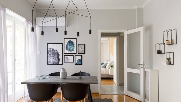 Uma área de refeições moderna com mesa, cadeiras e luminárias pendentes. Há obras de arte emolduradas na parede e uma porta que dá para outro cômodo.
