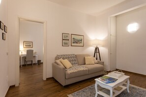 Una sala de estar acogedora con un sofá, una mesa de centro y un escritorio con una silla. Hay cuadros en la pared y una lámpara de pie.