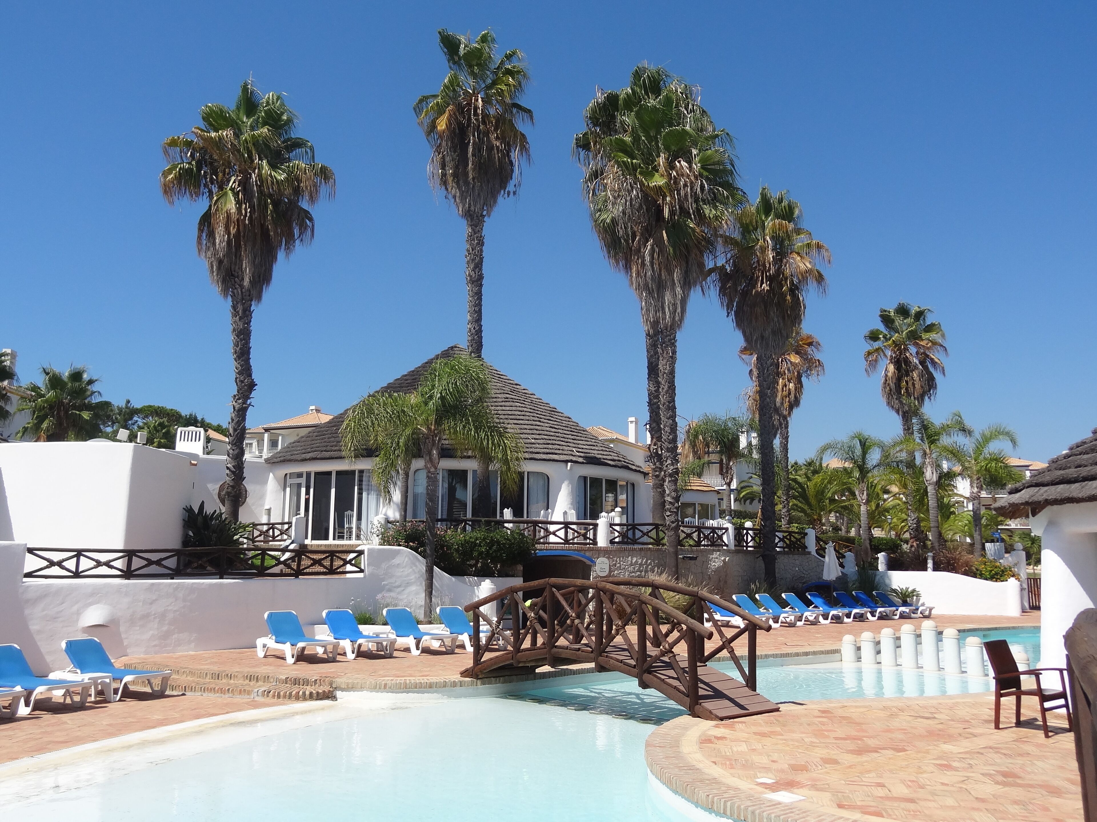 Zona de piscina amb gandules, palmeres i un edifici amb teulada enrajolada.