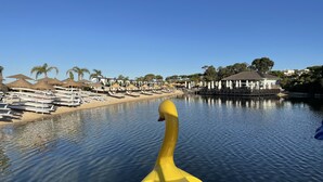 A waterfront area with lounge chairs and umbrellas, a building with a thatched roof, and a swan in the foreground. - Encosta do Lago  2 Bed Apartment with Private Garden, Large Palm Fringed Pool (Quinta do Lago)