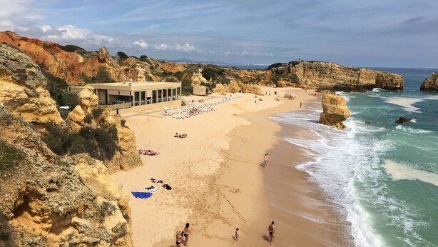 A coastal beach with rocky cliffs, a beachside building with outdoor seating, and clear skies. - Encosta do Lago  2 Bed Apartment with Private Garden, Large Palm Fringed Pool (Quinta do Lago)