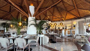 A spacious restaurant with high ceilings and a thatched roof. Tables are set with wine glasses and plates, and there are several large plants and hanging spherical lights. - Encosta do Lago  2 Bed Apartment with Private Garden, Large Palm Fringed Pool (Quinta do Lago)