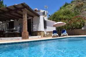 A pool area with two lounge chairs, a patio with a table and chairs, and a building with a tiled roof and stone pillar. - Romantica Finca The Hostel for 2 with private pool and fantastic views (Cómpeta)