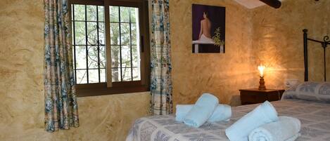 A bedroom with a bed, a nightstand, a window with curtains, a wall-mounted painting, and a lamp.