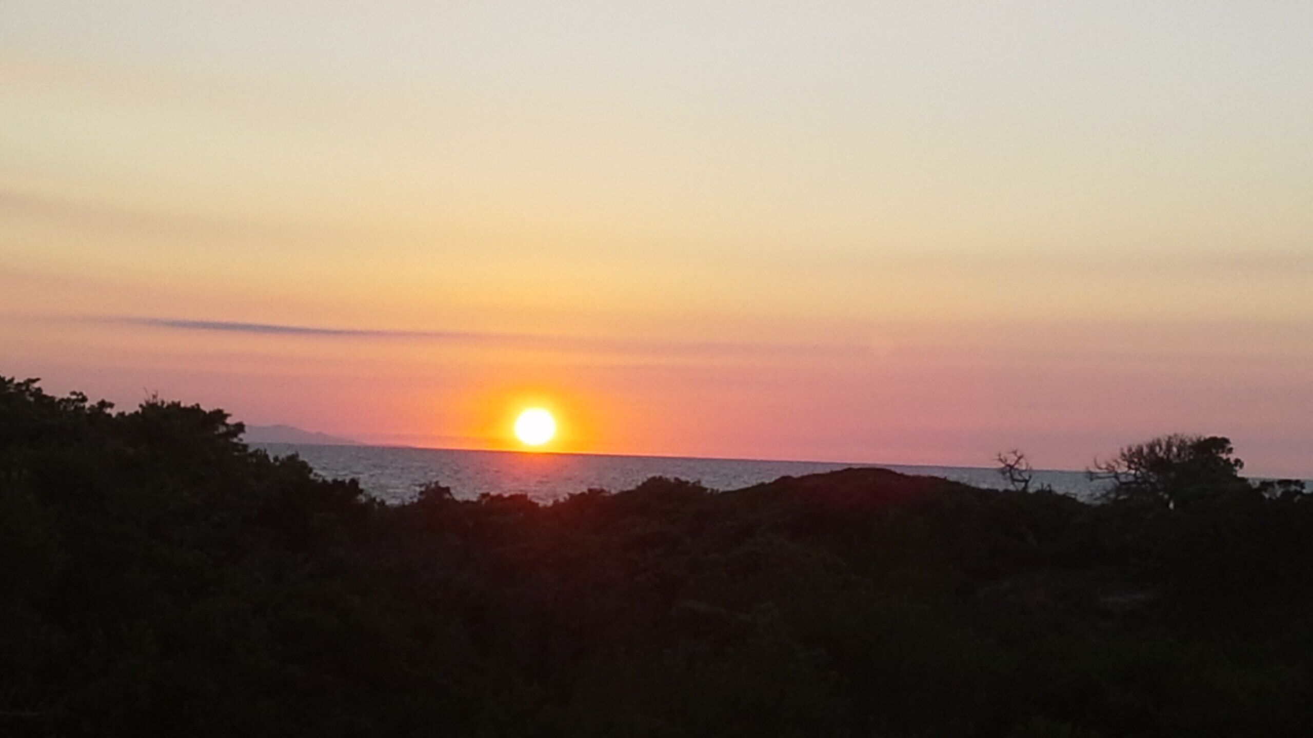 Ein Sonnenuntergang über einem Gewässer mit Baumreihen und einem fernen Gebirgszug im Hintergrund.