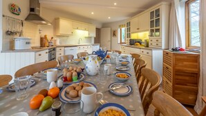 A well-lit kitchen with white cabinets, a dining table set with plates, bowls, and cups, and a variety of kitchen utensils hanging on the wall. - The Roundhouse has an unusual design and stands alone overlooking the lakes. (Wadebridge)