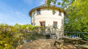 A round building with a wooden deck and picnic table, surrounded by greenery and trees. - The Roundhouse has an unusual design and stands alone overlooking the lakes. (Wadebridge)