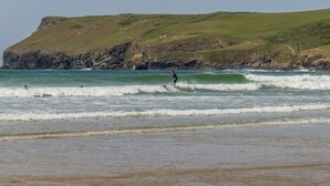 A coastal scene with a person surfing, a sandy beach, and a grassy hill in the background. - The Roundhouse has an unusual design and stands alone overlooking the lakes. (Wadebridge)