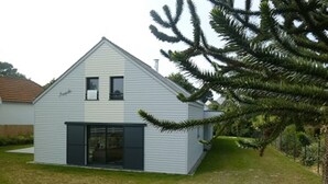 Une maison moderne à deux étages, dotée d’un toit en pente, d’un portail de garage à l’avant et d’un petit jardin avec un arbre.