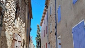 Una calle estrecha bordeada por edificios de piedra y enlucido, con contraventanas, un cielo despejado y un vehículo estacionado a lo lejos.