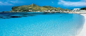 A coastal beach with clear blue waters, a sandy shore, and a mountain in the background.