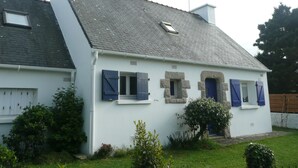 Una casa blanca con techo gris, contraventanas azules y un pequeño jardín.