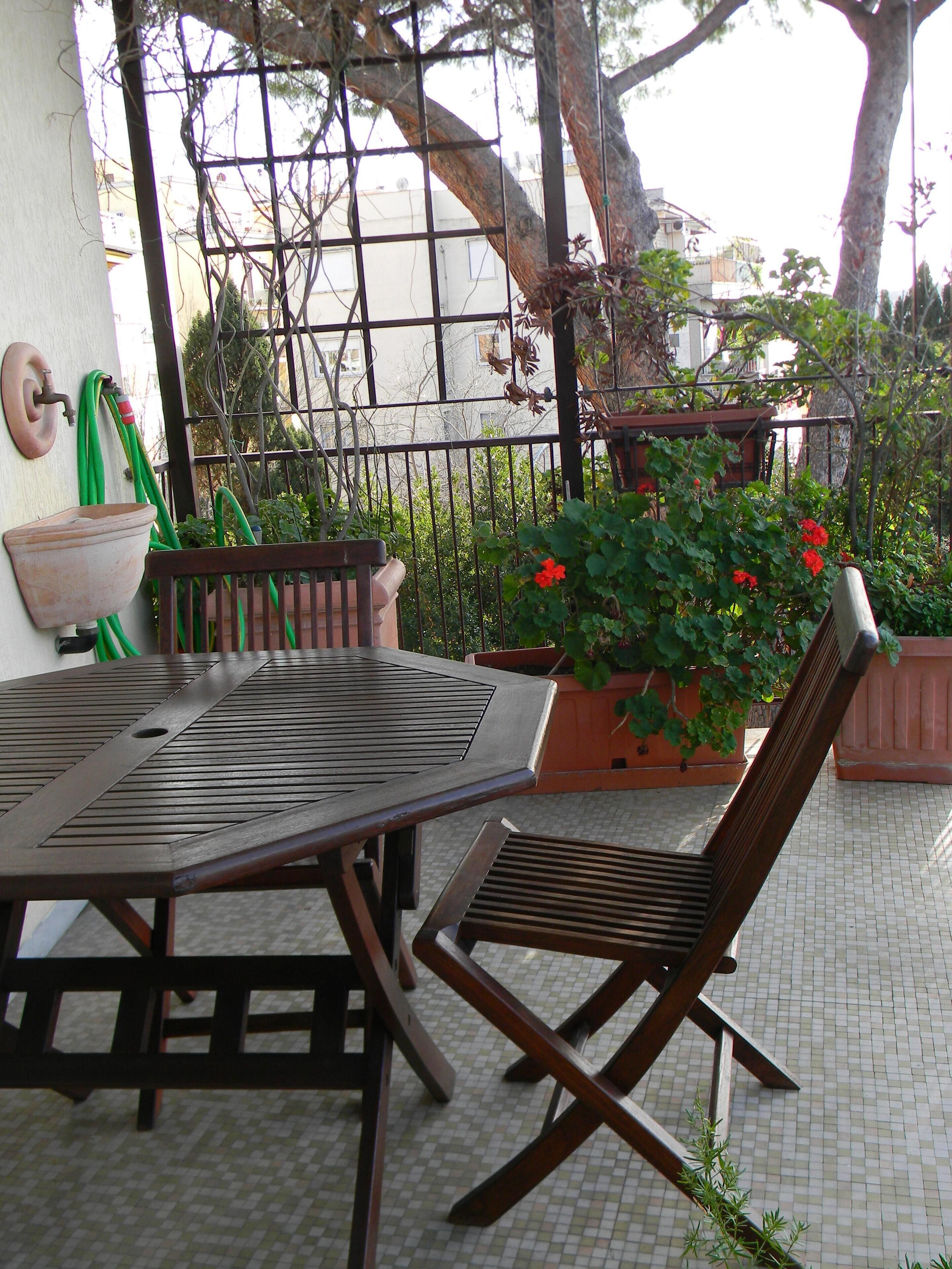 An outdoor patio with a table and chairs, surrounded by potted plants and a metal fence.