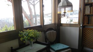 Une pièce avec vue sur le paysage urbain, grâce à de grandes fenêtres, un fauteuil avec un coussin, une petite table sur laquelle est posé un vase de fleurs et une bibliothèque.