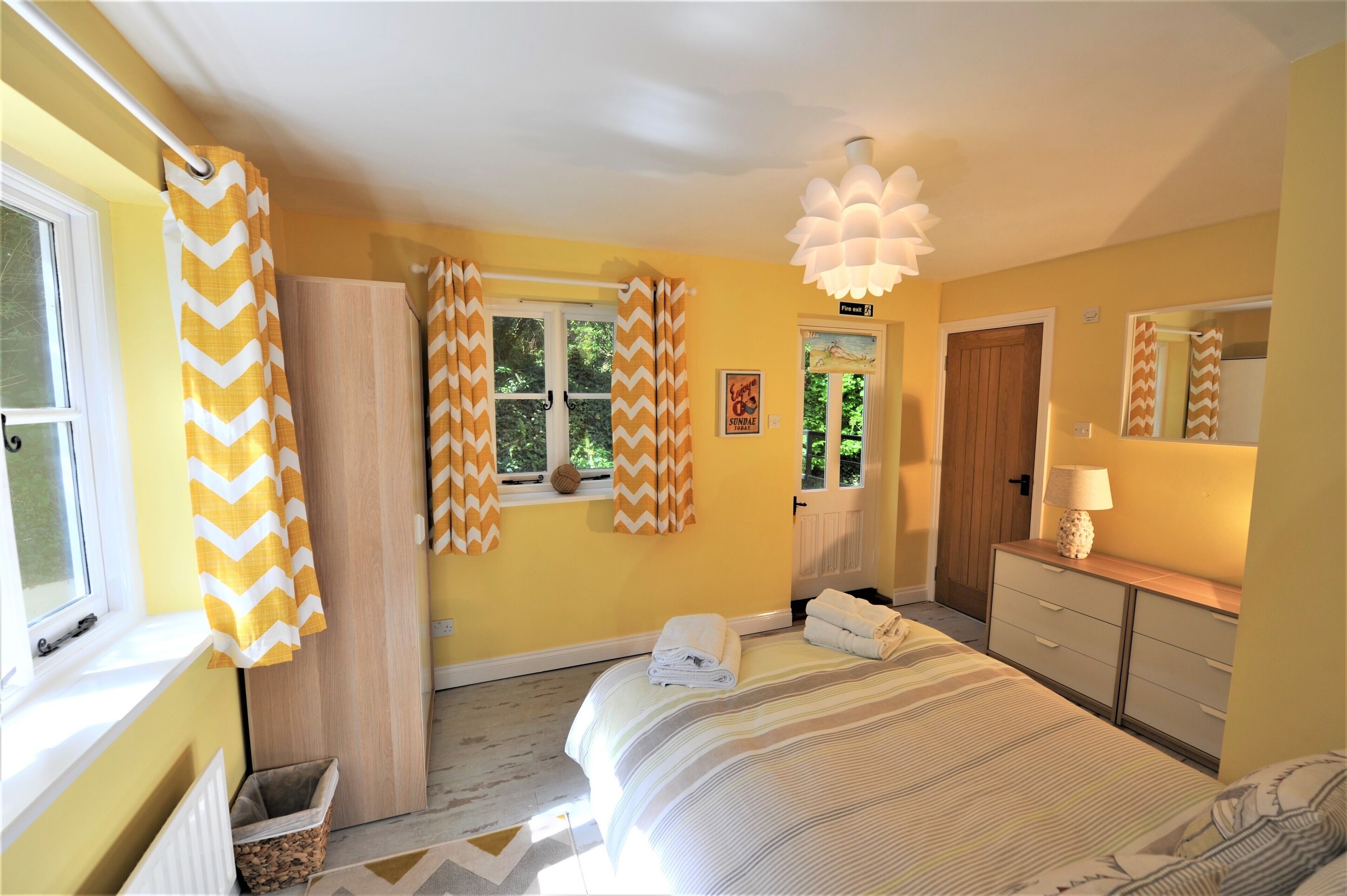 A bedroom with a bed, a bedside table with a lamp, a window with curtains, a radiator, and a wooden door.