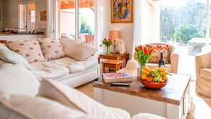 Un salon spacieux avec un canapé, une table basse sur laquelle se trouve un panier de fruits, un tableau accroché au mur et une vue sur l’extérieur grâce à de grandes fenêtres.
