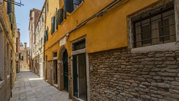 A narrow cobblestone street flanked by historic buildings with stone and plaster facades, arched doorways, and a mix of open and closed shutters.
