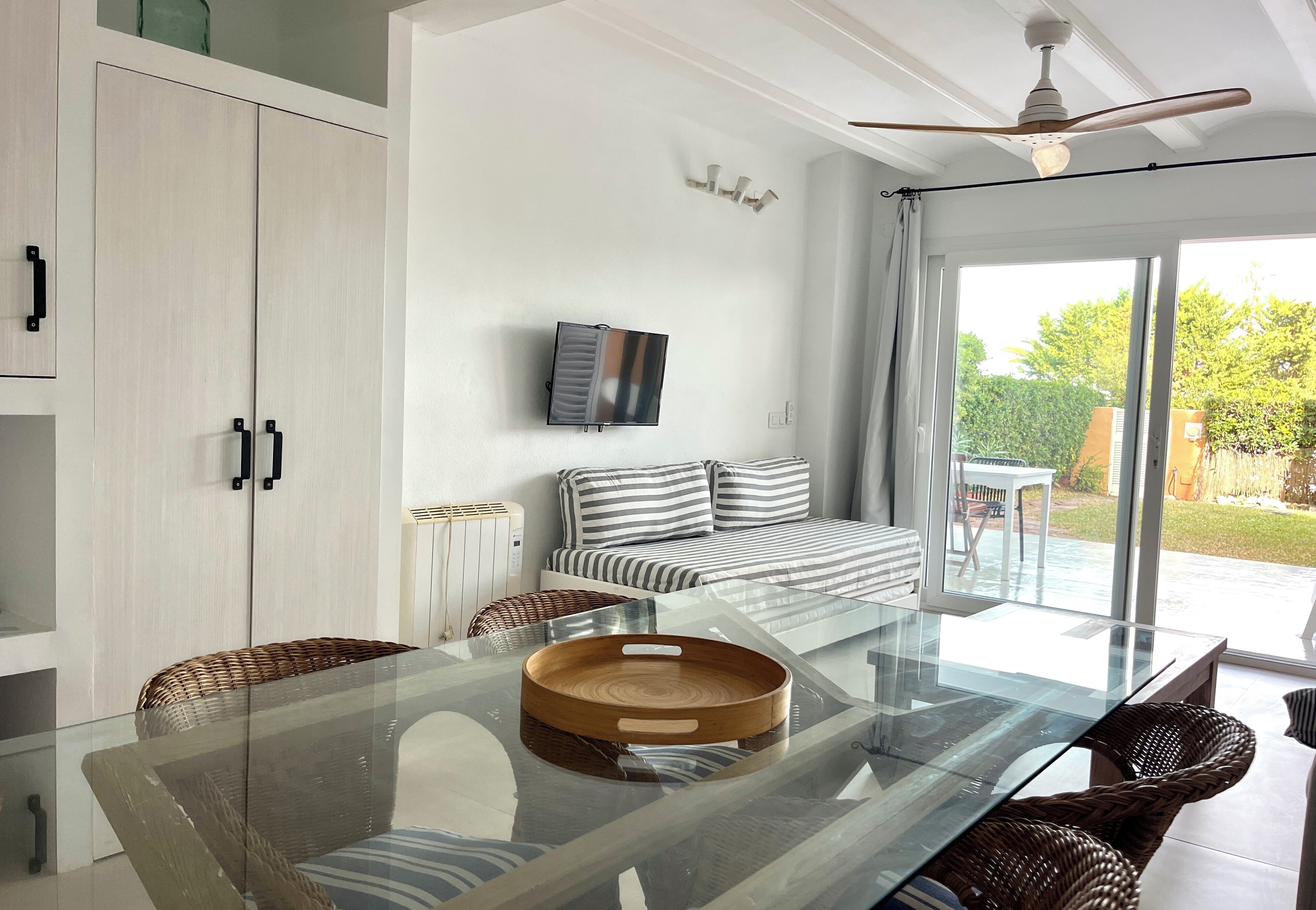 A modern living room with a glass dining table, wicker chairs, a sofa with striped cushions, and a television on the wall.