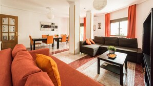 Ein modernes Wohnzimmer mit einer Eckcouch, einem Essbereich mit orangefarbenen Stühlen und einem großen Fenster mit orangefarbenen Vorhängen.