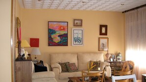 Ein Wohnzimmer mit einer Couch, Sesseln, einem Couchtisch und einem Esstisch. An den Wänden befinden sich Gemälde und auf dem Beistelltisch steht eine Lampe.