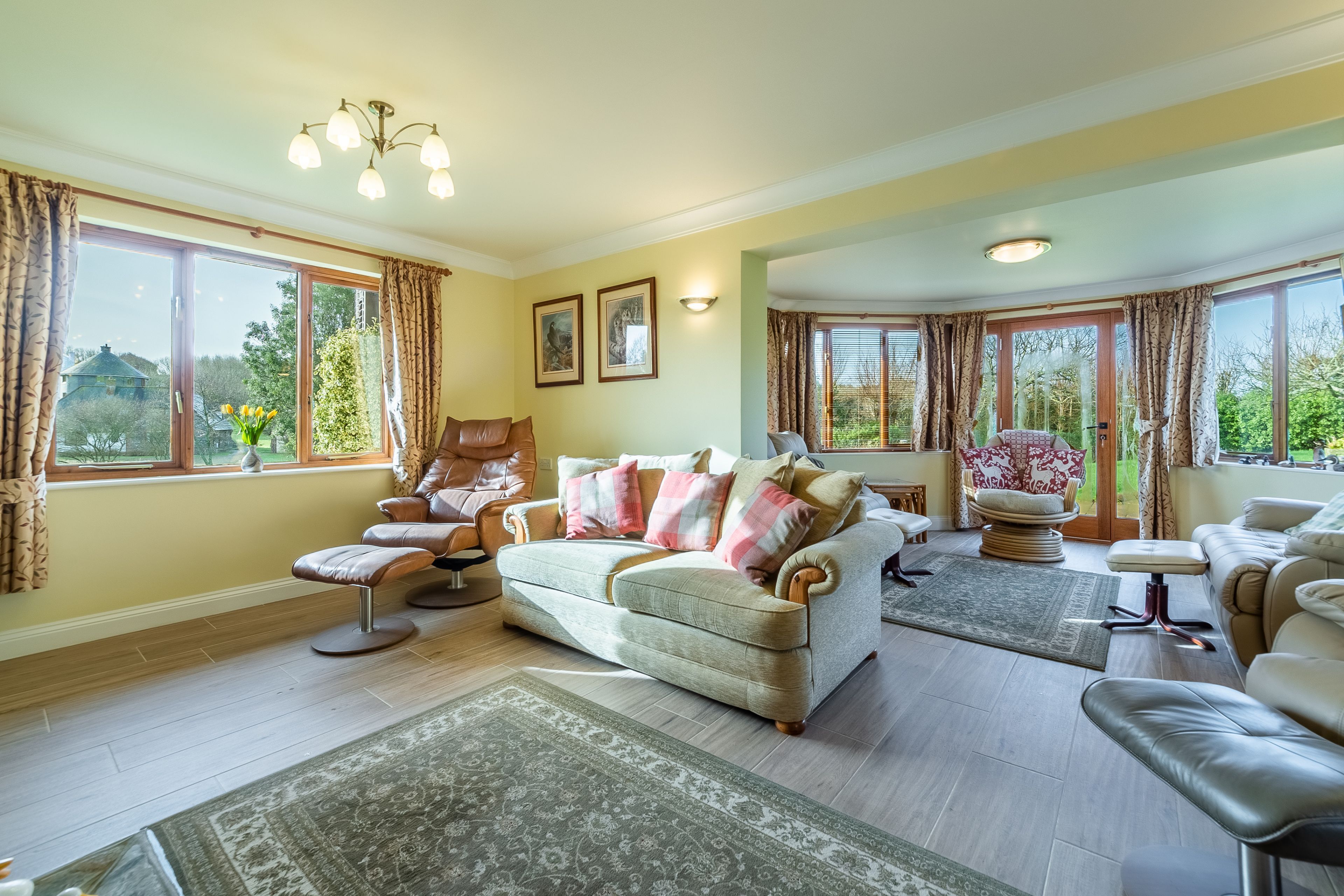A spacious living room with a beige sofa, leather armchairs, and a large window offering a view of the outdoors.