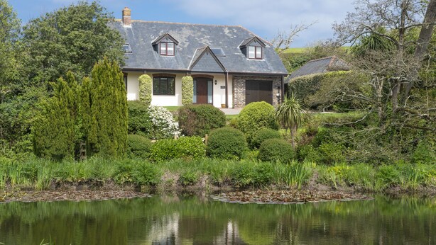 Une maison avec un étang devant, entourée d’une végétation luxuriante et d’arbres.