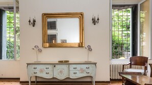 Ein Zimmer mit Holzboden, weißer Wand, großem Spiegel mit goldener Rahmung, weißem Schminktisch mit floralen Mustern, zwei Wandleuchten und einem Fenster mit Blick auf Grünflächen.