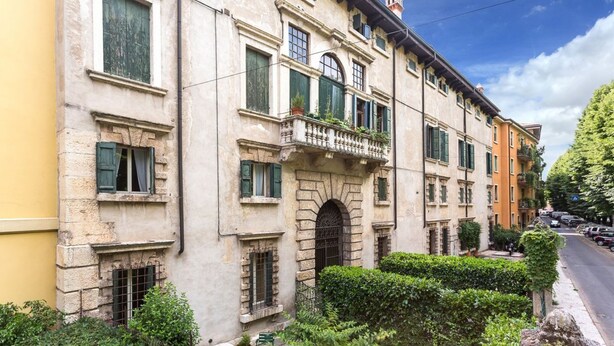 Ein historisches Gebäude mit einem zentralen Rundbogenportal, zahlreichen Fenstern mit Fensterläden und einem kleinen Balkon mit Blumentöpfen.