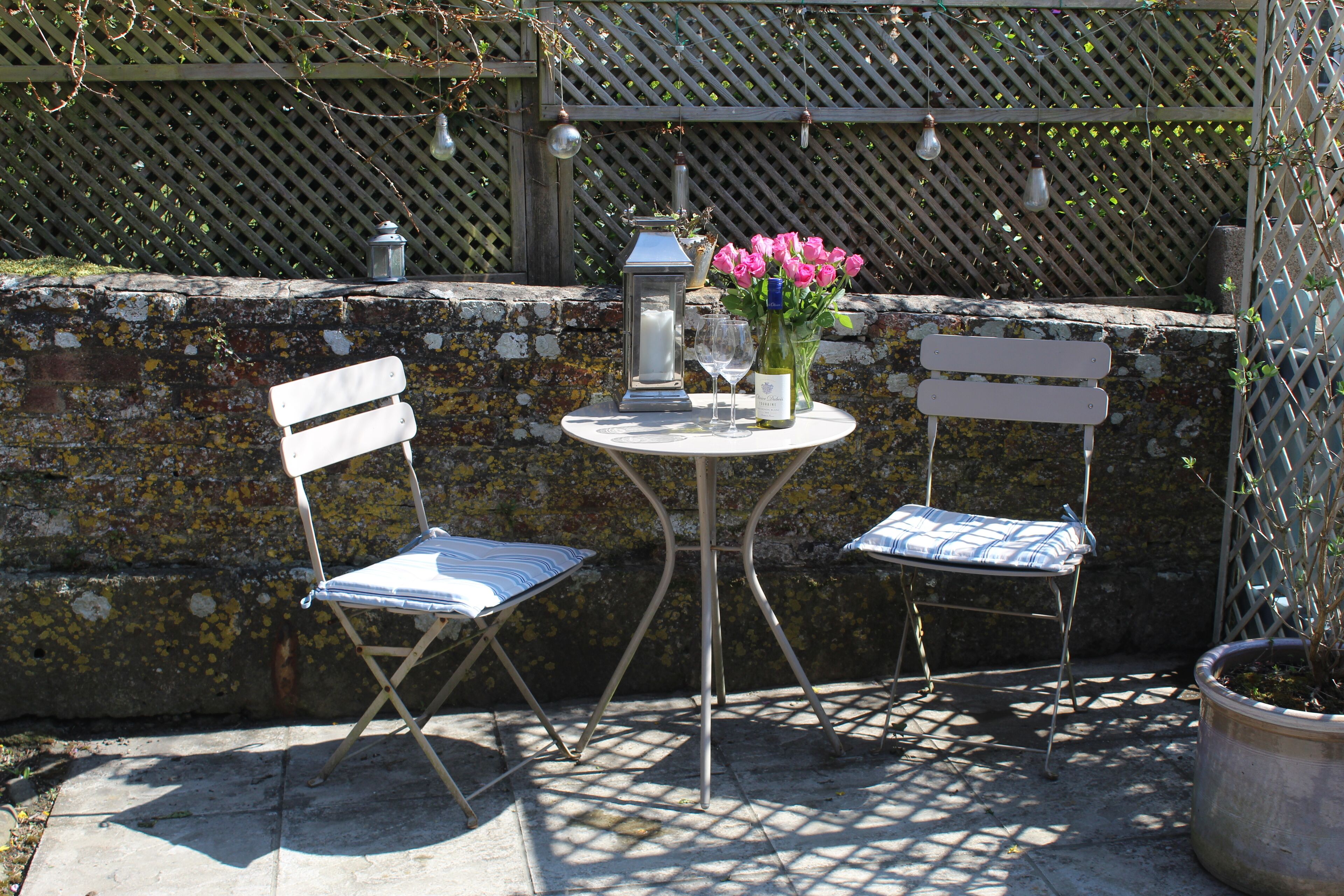 An outdoor seating area with two white folding chairs, a small round table, a candle, a wine glass, and a vase with pink flowers.