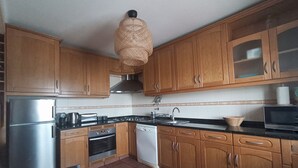 A kitchen with wooden cabinets, stainless steel appliances, and a hanging light fixture. - Holiday Home in Sines (Sines)