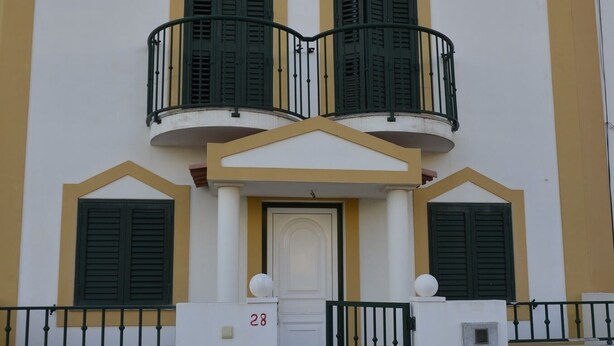 A two-story building with a white facade and yellow trim, featuring a central entrance with a white door and green shutters, and a balcony with black railings. - Holiday Home in Sines (Sines)